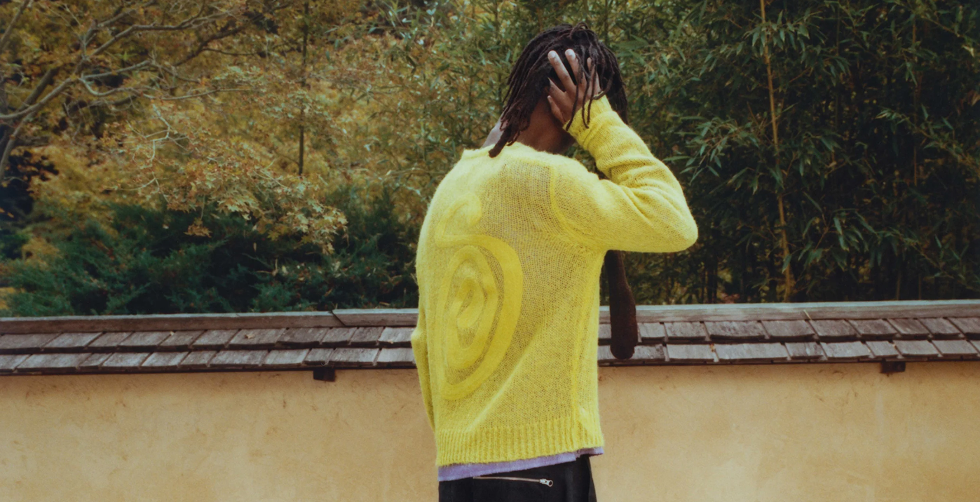 Person in a yellow sweater standing with their back to the camera, hand on head, in front of a lush green backdrop.